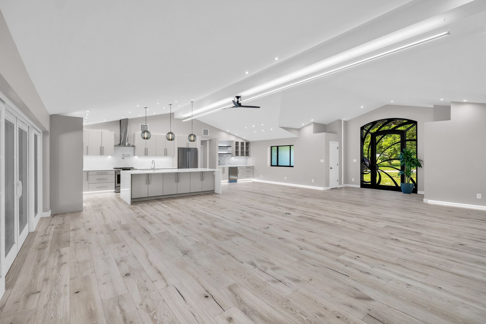 Open great room and kitchen with a vaulted ceiling, linear LED lighting, wide-plank light oak floors, a white waterfall-edge island, and an arched black iron front door